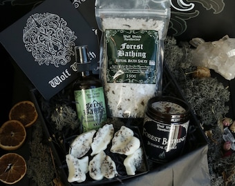 May include: A black gift box filled with a forest-themed bath and body set. The set includes a bottle of body wash, a bag of bath salts, and a candle. All items feature a green and white label with the text "Forest Bathing" and "Wolf Wytch Apothecary".