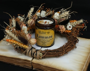 May include: A brown glass candle jar with a gold label that reads "Wolf Witch Apothecary Lammas Grass Patchouli & Hemlock Coconut Oat candle 180ml Net Weight". The candle is surrounded by a dried floral wreath with wheat stalks, dried flowers, and seed pods.