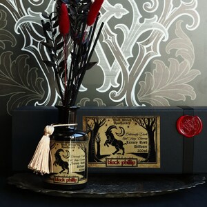 Könnte beinhalten: Eine schwarze Glasflasche mit einem goldenen Etikett und einem schwarzen Quasten. Das Etikett zeigt eine schwarze Ziege mit einem Halbmond und dem Text "Wolf Wytch Apothecary" und "Deliciously Dark Red Juicy Cherries Luxury Reed Diffuser 200ml". Die Flasche steht auf einer schwarzen Box mit einem roten Wachssiegel.