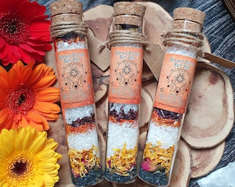 May include: Three glass vials filled with layered bath salts, herbs, and flowers. Each vial has a cork stopper and a brown paper label that reads "Summer Solstice Bath Shot".