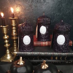 May include: Four black pillar candles with white wax medallions on a stack of books. The candles are on a dark surface with two gold candle holders.