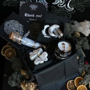 May include: A black gift box filled with various items, including a candle, bath bombs, and a small glass tube. The box is surrounded by dried flowers and leaves. The candle is labeled "White Sage & Rose Quartz" and "Mama Magic Botanicals".