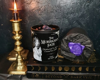 May include: Black candle jar with a black and gold label that reads "The Morrigan's Fate Blackcurrent & Coconut wax candle 100g Net Weight". The candle is sitting on a stack of books with a black cover and gold lettering that reads "Beyond the Dark Veil".