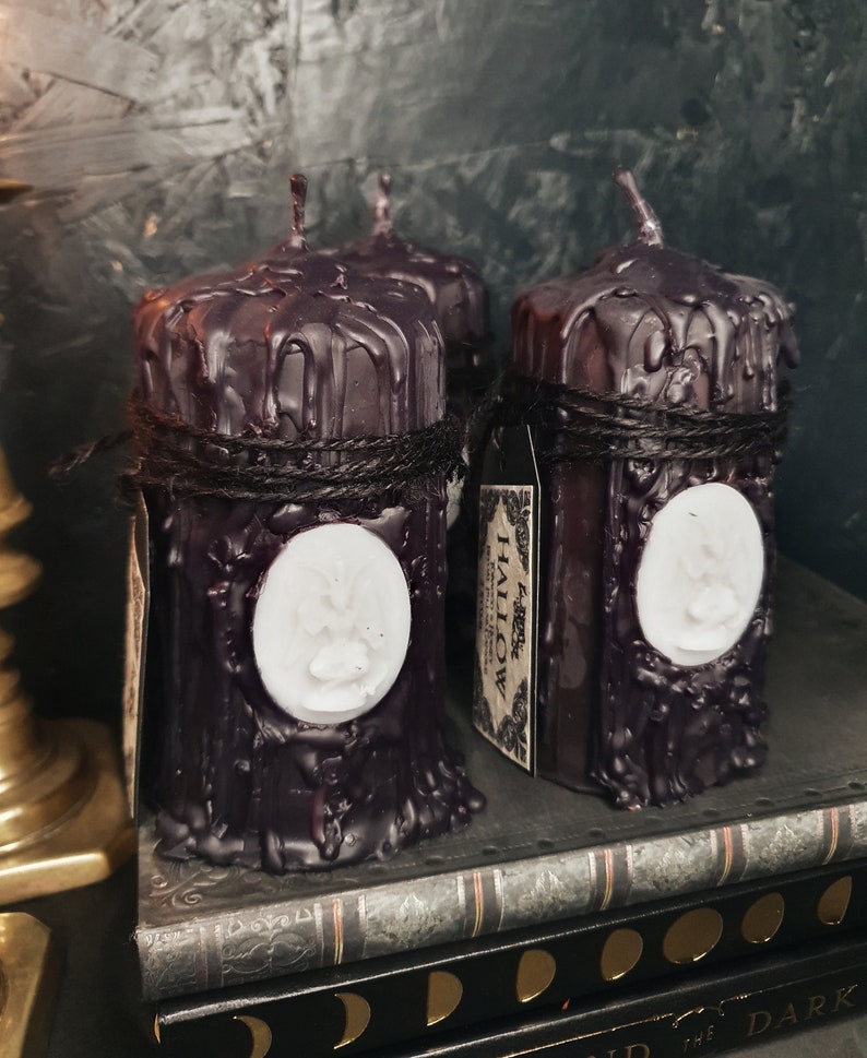 May include: Two black pillar candles with a white wax design of a bat on the front. The candles are wrapped with twine and have a label that reads "Forest Hallow".