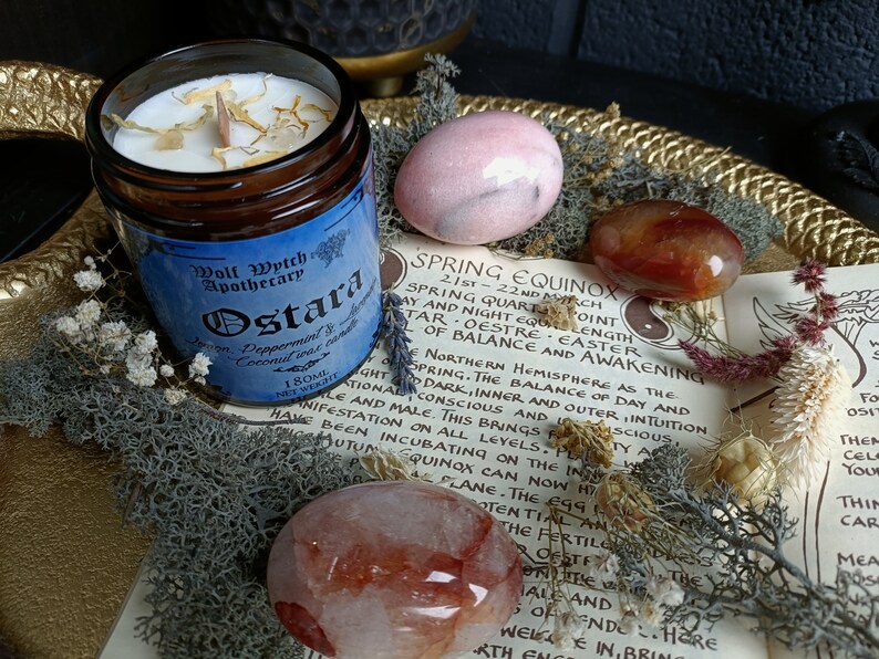 May include: A blue and gold candle jar with the label "Wolf Wytch Apothecary Ostara" and the text "Lemon, Peppermint & Coconut incense candle 180ml Net Weight". The candle jar is surrounded by pink and red stones, dried flowers, and moss.