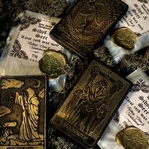 May include: Four tarot card wax melts with gold foil accents and wax seals. The cards are The Hermit, The Empress, The World, and Sibyl Seer. The wax melts are packaged in white paper with black text and are labeled with the card name, scent, and weight.