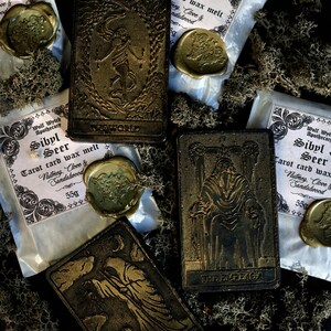 May include: Four tarot cards with gold accents and the words "The World", "The Hermit", "The Emperor", and "The Empress" on them. The cards are surrounded by small bags of wax melt with the text "Sibyl & Seer Tarot card wax melt" and the scents "Nutmeg, Clove & Sandalwood".