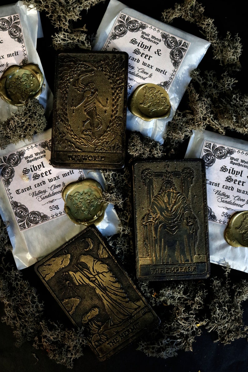 May include: Four tarot cards with gold accents and the words "The World", "The Hermit", "The Emperor", and "The Empress" on them. The cards are surrounded by small bags of wax melt with the text "Sibyl & Seer Tarot card wax melt" and the scents "Nutmeg, Clove & Sandalwood".