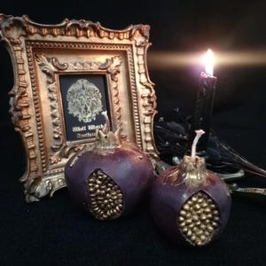 May include: Two purple pomegranate-shaped candles with gold detailing. The candles are sitting on a black surface in front of a gold picture frame with a black and white image inside.
