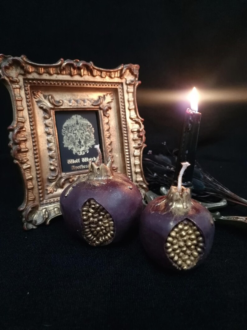 May include: Two purple pomegranate-shaped candles with gold detailing. The candles are sitting on a black surface in front of a gold picture frame with a black and white image inside.