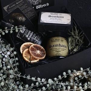 May include: A black gift box filled with a variety of items, including a candle, a bottle of essential oil, and dried orange slices. The candle is labeled "Wolf Watch Apothecary Samhain Wild Orange Clove & Ginger Coconut Wax Candle 180ml Net Weight". The essential oil bottle is labeled "Sacred Samhain Crystal Infused Ritual Sanctums 100ml Handmade". The box is decorated with a small tag that says "Wytch".