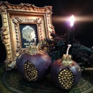 May include: Two purple pomegranate-shaped candles with gold detailing on a gold tray. The candles are lit and the flames are visible. A gold framed picture with a black background and gold design is in the background.