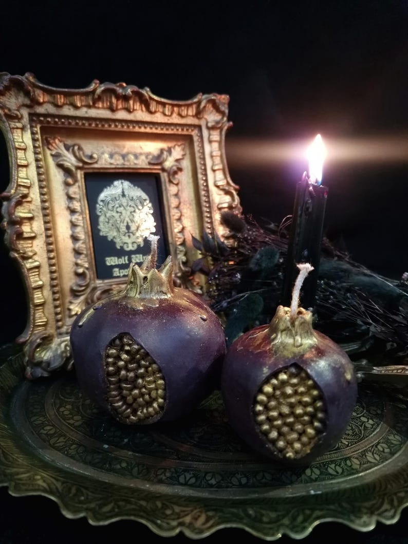 May include: Two purple pomegranate-shaped candles with gold detailing on a gold tray. The candles are lit and the flames are visible. A gold framed picture with a black background and gold design is in the background.