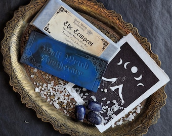 May include: A still-life arrangement featuring a gold-toned tray with a blue bar of soap, a wrapped bar labeled "The Tempest," and a black and white print with moon phases. Scattered white crystals and blue stones add detail.