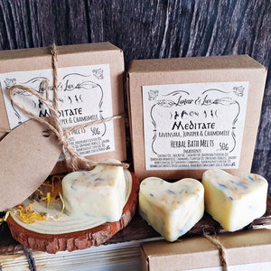 May include: Two boxes of herbal bath melts, each with a heart-shaped melt. The boxes are brown with a label that reads "Meditate" and "Juniper & Chamomile". The melts are off-white with visible herbs.