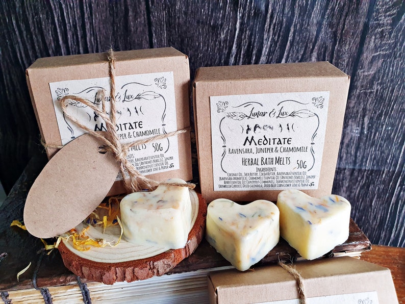 May include: Two boxes of herbal bath melts, each with a heart-shaped melt. The boxes are brown with a label that reads "Meditate" and "Juniper & Chamomile". The melts are off-white with visible herbs.