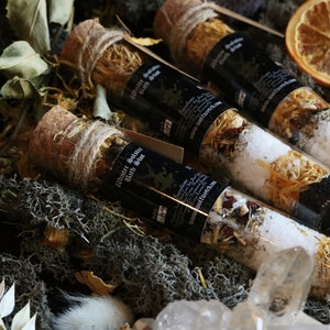 May include: Three glass vials filled with bath salts and dried herbs, tied with twine and corked. The vials are labeled "Winter Solstice Bath Shot" and have a website address. The vials are arranged on a bed of moss and crystals.