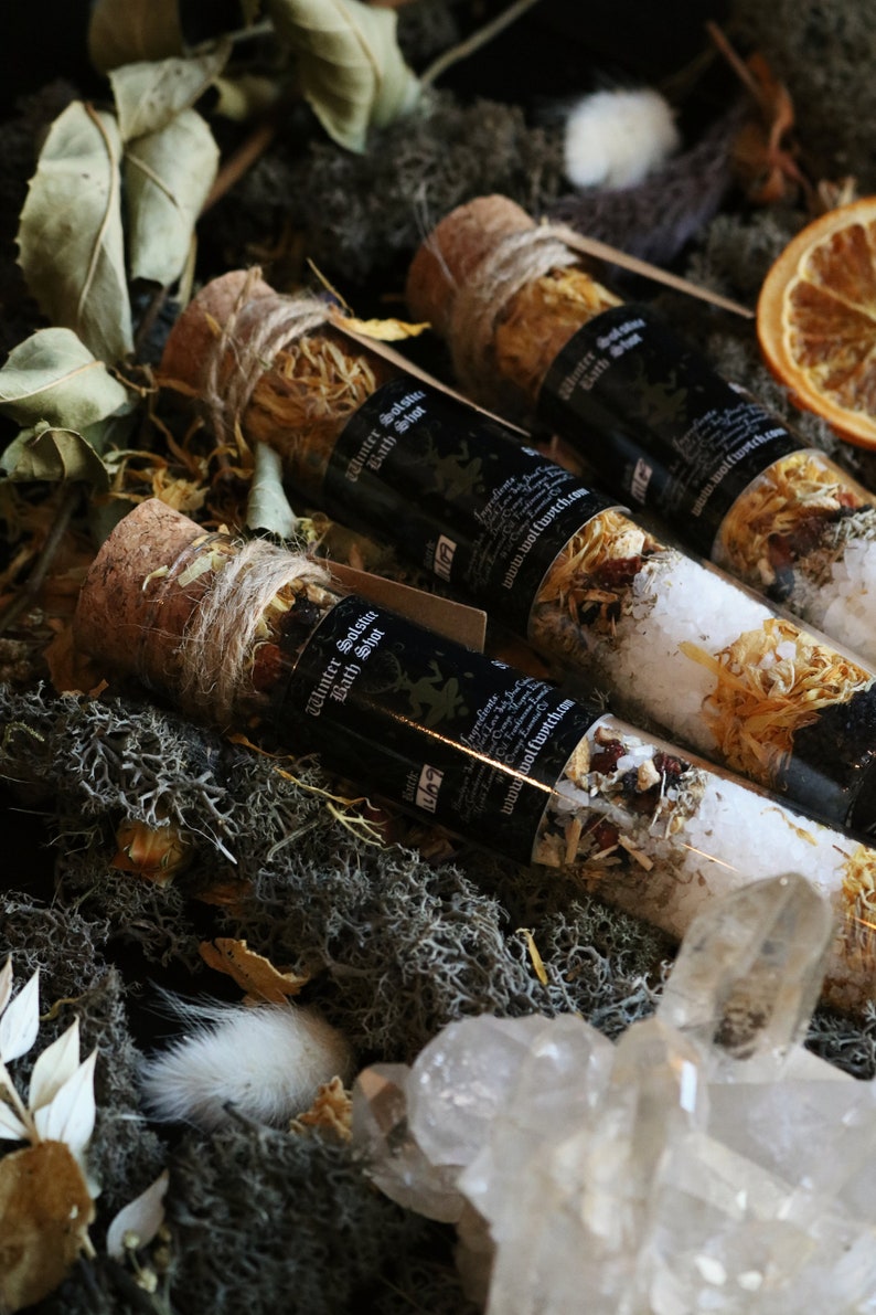 May include: Three glass vials filled with bath salts and dried herbs, tied with twine and corked. The vials are labeled "Winter Solstice Bath Shot" and have a website address. The vials are arranged on a bed of moss and crystals.