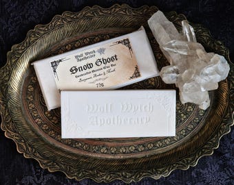 May include: A decorative gold-toned tray holds two white wax bars and a clear quartz crystal. One bar is labelled "Snow Ghost" and "Wolf Witch Apothecary." The other bar is embossed with "Wolf Witch Apothecary." The label reads 75g.