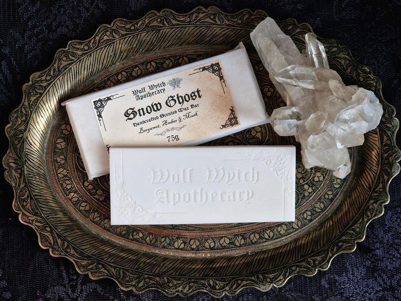 K&ouml;nnte beinhalten: Zwei wei&szlig;e Wachsriegel und ein klarer Bergkristall liegen auf einem dekorativen Tablett in Goldt&ouml;nen. Ein Riegel ist mit "Snow Ghost" und "Wolf Witch Apothecary" beschriftet. Der andere Riegel ist mit "Wolf Witch Apothecary" gepr&auml;gt.