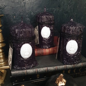 May include: Three black pillar candles with a white wax design on the front. The candles are sitting on a stack of books with a black cover. The candles are decorated with black twine.
