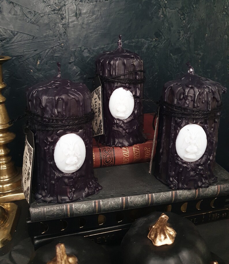 May include: Three black pillar candles with a white wax design on the front. The candles are sitting on a stack of books with a black cover. The candles are decorated with black twine.