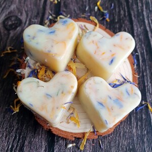 May include: Four heart-shaped wax melts with floral inclusions, resting on a small wooden slice. The melts are creamy white with blue, yellow, and orange petals. The background is a dark wood surface, with scattered petals.