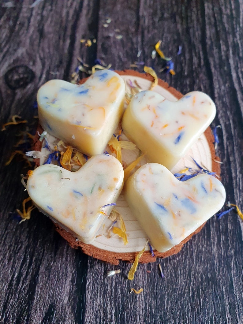 May include: Four heart-shaped wax melts with floral inclusions, resting on a small wooden slice. The melts are creamy white with blue, yellow, and orange petals. The background is a dark wood surface, with scattered petals.