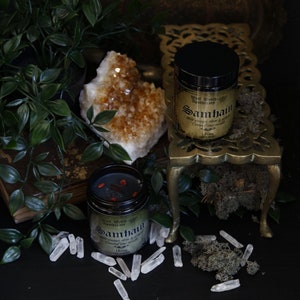 May include: Two glass jars with black lids and labels. The jars contain a dark red liquid and are labeled "Samhain" with a witch's apothecary logo. The jars are on a gold metal stand with a crystal cluster and green foliage in the background.
