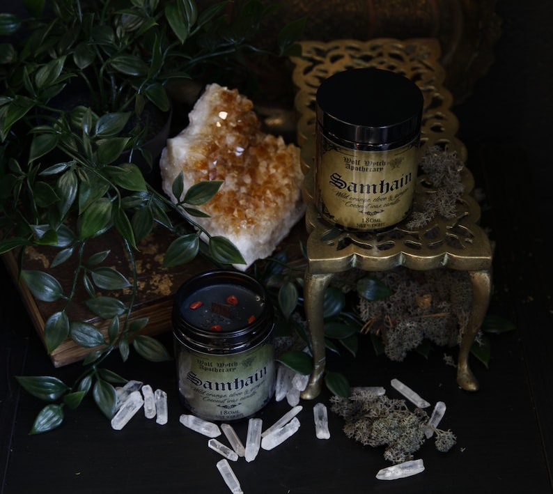 May include: Two glass jars with black lids and labels. The jars contain a dark red liquid and are labeled "Samhain" with a witch's apothecary logo. The jars are on a gold metal stand with a crystal cluster and green foliage in the background.