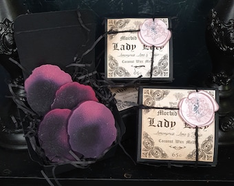 May include: A black gift box containing purple and pink floral wax melts, alongside two black boxes of "Morbid Lady Lavy" coconut wax melts, each sealed with a pink wax stamp and tied with black twine. The wax melts are 65g.