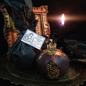 May include: A black and gold pomegranate-shaped candle on a gold tray. The candle is surrounded by black fabric and a black candle in a glass holder. The candle has the text "Whole Spices & Aromatherapy - PERSEPHONE'S FRUIT -  Black Raspberry, Pomegranate, Patchouli, Musk - 270g" on a label.