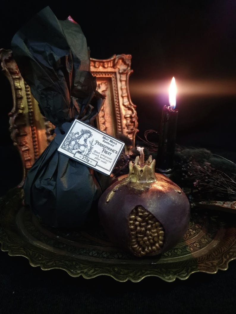 May include: A black and gold pomegranate-shaped candle on a gold tray. The candle is surrounded by black fabric and a black candle in a glass holder. The candle has the text "Whole Spices & Aromatherapy - PERSEPHONE'S FRUIT -  Black Raspberry, Pomegranate, Patchouli, Musk - 270g" on a label.