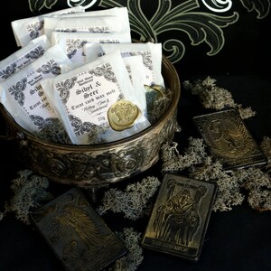 May include: A collection of white paper packets with a gold wax seal, labeled "Sibyl & Seer Tarot card wax melt" with the scent "Nutmeg, Clove & Sandalwood" and a weight of 55g. The packets are in a silver bowl with a decorative design. The bowl is sitting on a black surface with a few gold tarot cards scattered around it.