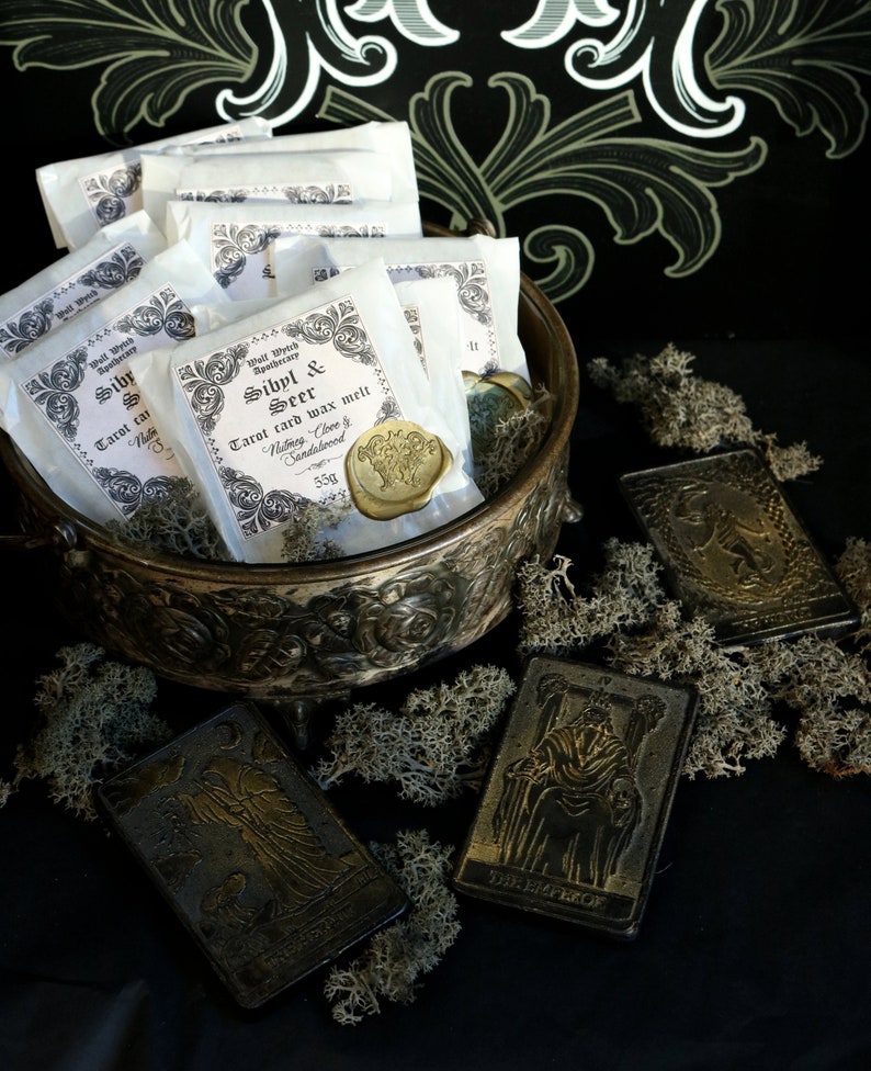 May include: A collection of white paper packets with a gold wax seal, labeled "Sibyl & Seer Tarot card wax melt" with the scent "Nutmeg, Clove & Sandalwood" and a weight of 55g. The packets are in a silver bowl with a decorative design. The bowl is sitting on a black surface with a few gold tarot cards scattered around it.