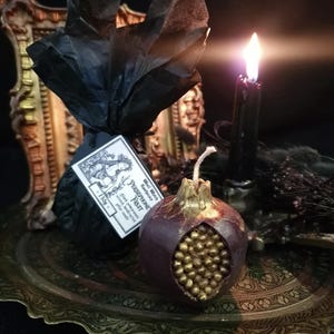 May include: A dark red pomegranate-shaped candle with gold details. The candle is sitting on a gold tray with a black candle and a small black bag with a label that reads "Black Moon Apothecary - Persephone Fruit - Hand poured soy wax with essential oils - 11.5 oz."