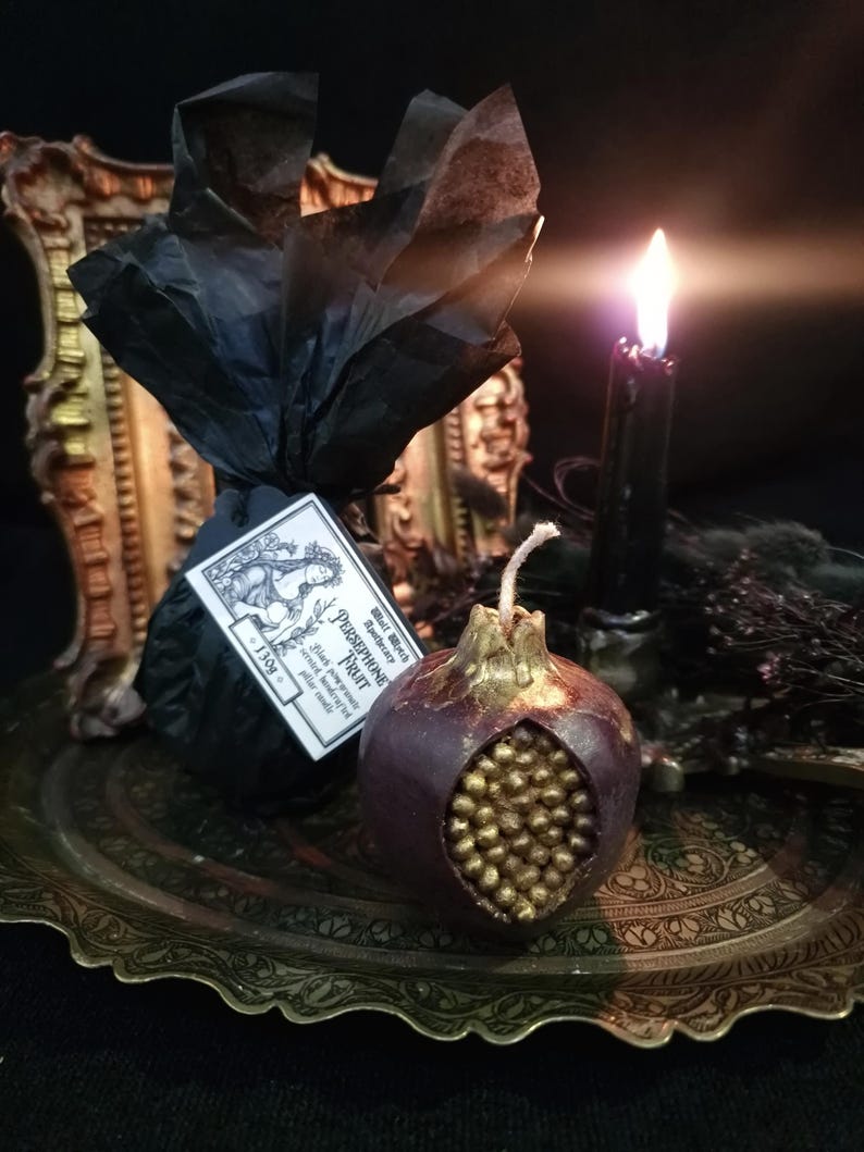 May include: A dark red pomegranate-shaped candle with gold details. The candle is sitting on a gold tray with a black candle and a small black bag with a label that reads "Black Moon Apothecary - Persephone Fruit - Hand poured soy wax with essential oils - 11.5 oz."
