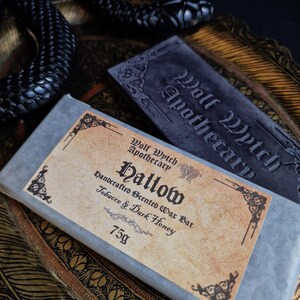 May include: A close-up of a handcrafted scented wax bar labeled "Hallow" by Wolf Wytch Apothecary. The bar is wrapped in gray paper with a label that reads "Tobacco & Dark Honey" and weighs 75g. A black snake and a gray stone with the words "Wolf Wytch Apothecary" are also visible.