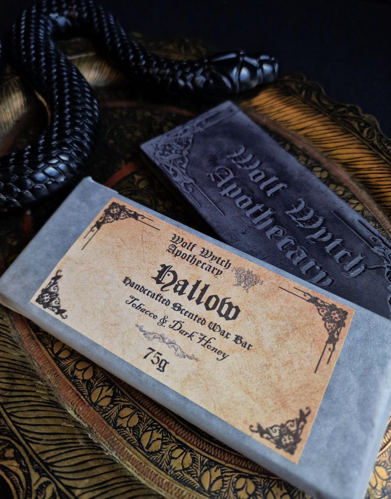 May include: A close-up of a handcrafted scented wax bar labeled "Hallow" by Wolf Wytch Apothecary. The bar is wrapped in gray paper with a label that reads "Tobacco & Dark Honey" and weighs 75g. A black snake and a gray stone with the words "Wolf Wytch Apothecary" are also visible.