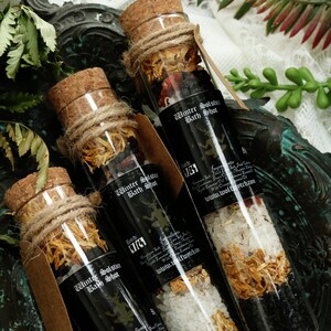 May include: Three glass bottles filled with bath salts and dried flowers. The bottles are labeled "Winter Solstice Bath Shot" and have a black background with white text. The bottles are sitting on a dark green surface.