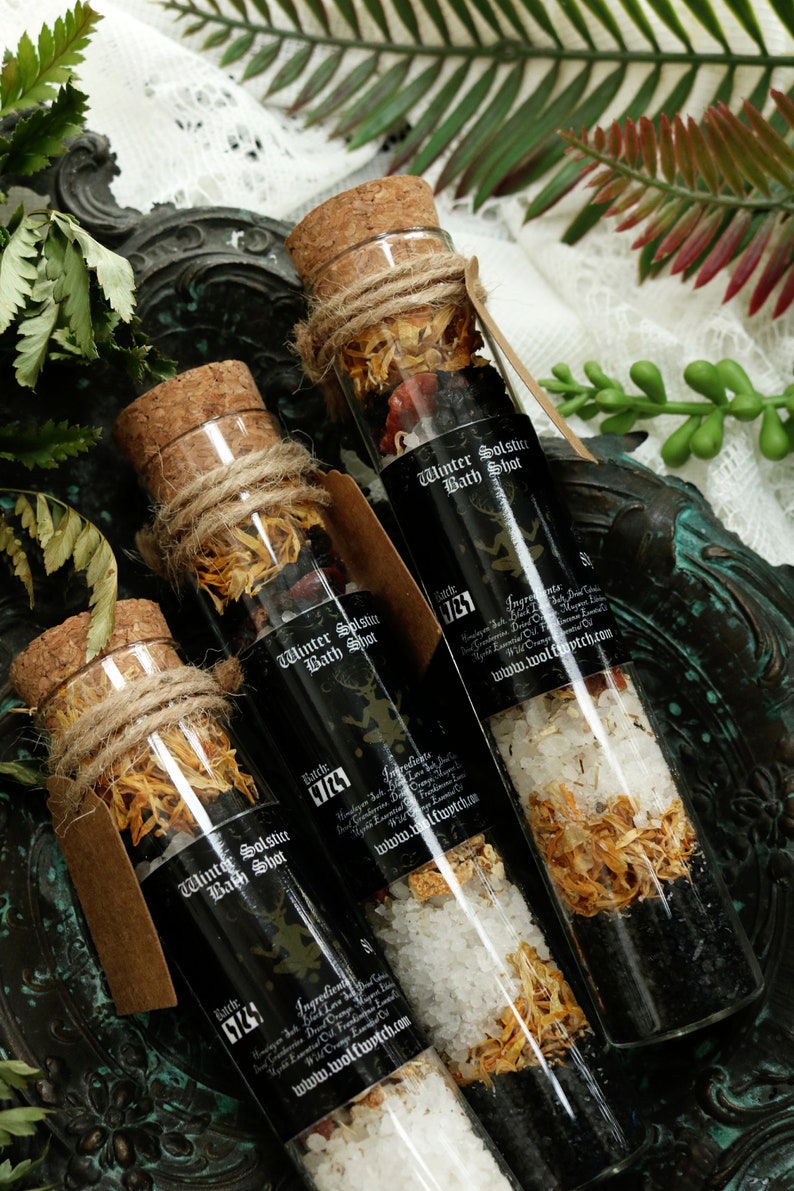 May include: Three glass bottles filled with bath salts and dried flowers. The bottles are labeled "Winter Solstice Bath Shot" and have a black background with white text. The bottles are sitting on a dark green surface.
