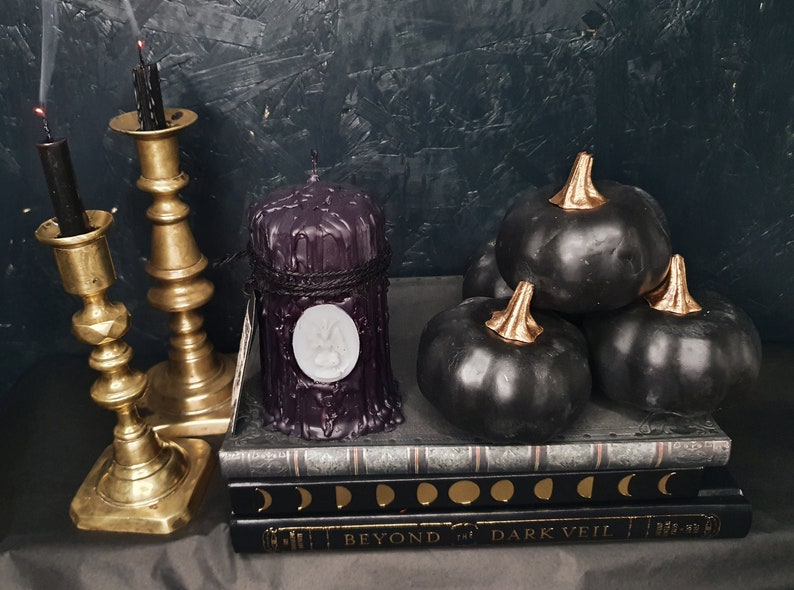 May include: A black pillar candle with a white wax seal sits on a stack of books titled "Beyond the Dark Veil". The candle is surrounded by black pumpkins with gold accents.