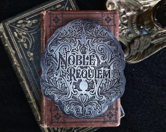 Peut inclure: Un autocollant en métal argenté avec un motif orné et les mots "NOBLE REQUIEM" dans une police gothique.