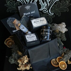 Op de afbeelding: Een zwarte cadeaudoos gevuld met heksenartikelen, waaronder een kaars, badzout en een bedankkaart. De doos is versierd met gedroogde sinaasappelschijfjes en mos.