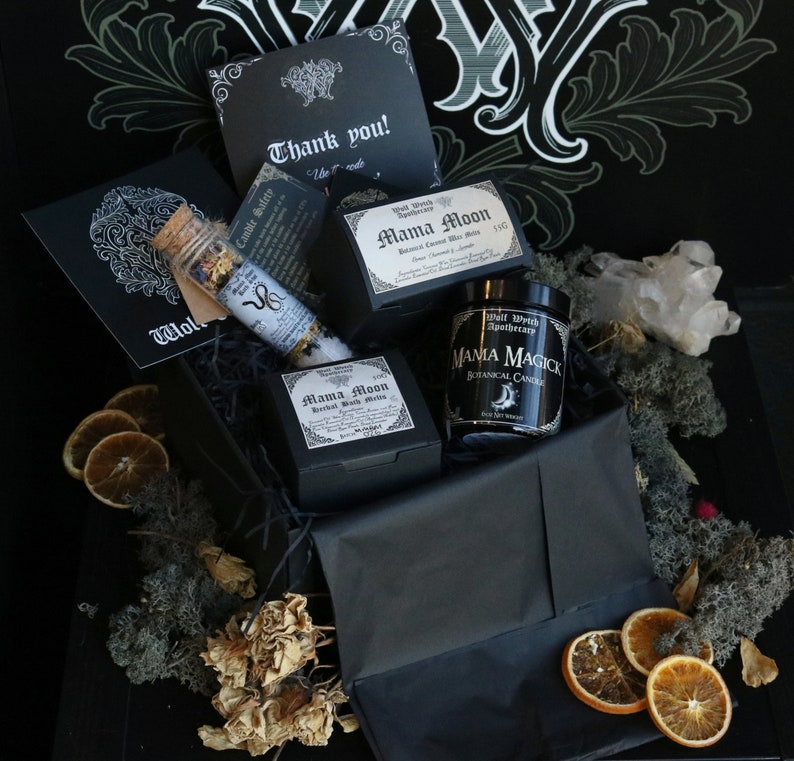 Op de afbeelding: Een zwarte cadeaudoos gevuld met heksenartikelen, waaronder een kaars, badzout en een bedankkaart. De doos is versierd met gedroogde sinaasappelschijfjes en mos.