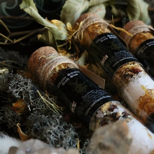 May include: Three small glass bottles with cork tops filled with bath salts and tied with twine. The bottles are labeled "Winter Solstice Bath Shot" and have a black and white label with a wolf design. The bottles are sitting on a bed of moss and dried leaves.