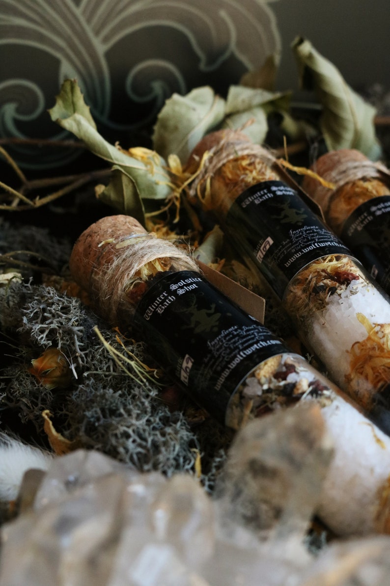 May include: Three small glass bottles with cork tops filled with bath salts and tied with twine. The bottles are labeled "Winter Solstice Bath Shot" and have a black and white label with a wolf design. The bottles are sitting on a bed of moss and dried leaves.