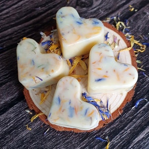 May include: Four heart-shaped wax melts with embedded blue, yellow, and orange flower petals rest on a wooden slice. The melts are creamy white, and scattered petals surround the base, creating a natural, floral aesthetic.