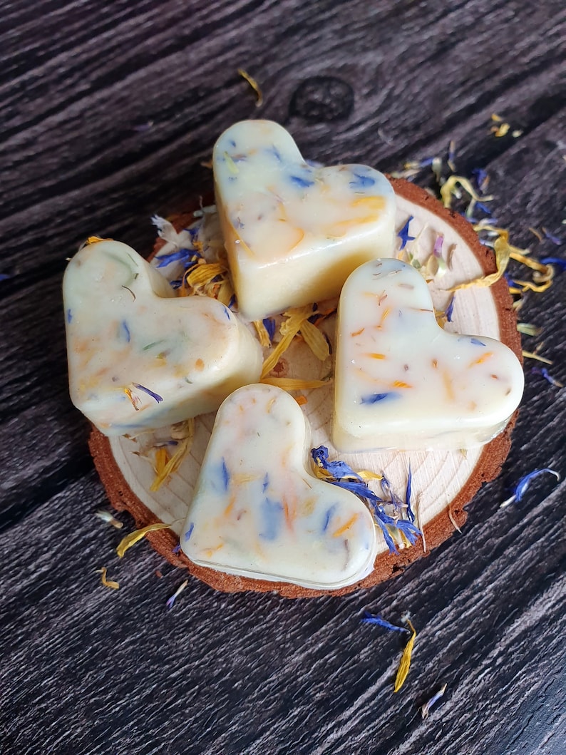 May include: Four heart-shaped wax melts with embedded blue, yellow, and orange flower petals rest on a wooden slice. The melts are creamy white, and scattered petals surround the base, creating a natural, floral aesthetic.