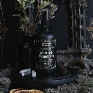 May include: A black glass bottle with a spray nozzle, labeled "Wolf Wythch Apothecary" and "Sacred Samhain Crystal Infused Ritual Saining Spray 100ML Handmade in the UK". The bottle is surrounded by dried orange slices, clear quartz crystals, and green moss.